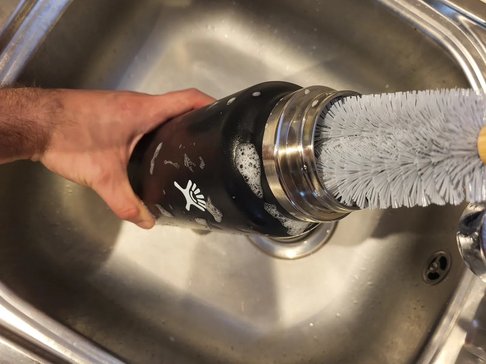 A hand cleaning the inside of a black water bottle with a bottle brush over a sink.