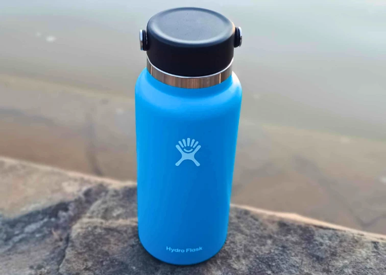 A 32 oz. blue Hydro Flask water bottle with a black cap, standing next to the water with the logo visible.