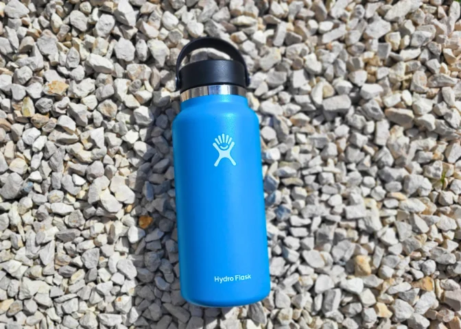 Blue 32 oz Hydro Flask water bottle with black cap on gravel, showing logo and brand name on stainless steel body.