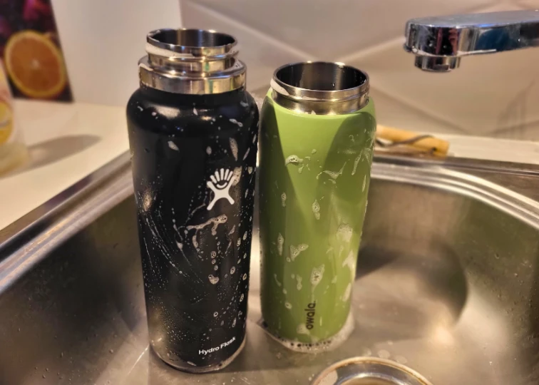 A Hydro Flask and Owala FreeSip water bottles are being cleaned in a sink with soap suds on them.

