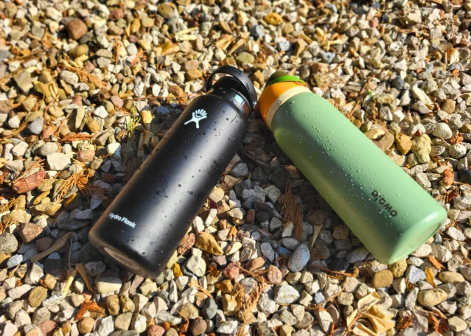 Two water bottles, a black Hydro Flask and a green Owala FreeSip, lie side by side on a gravel surface.