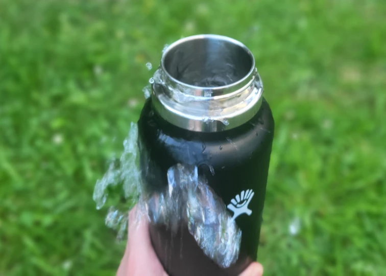 A black Hydro Flask with a wide mouth, water splashing out, against a blurred green background.