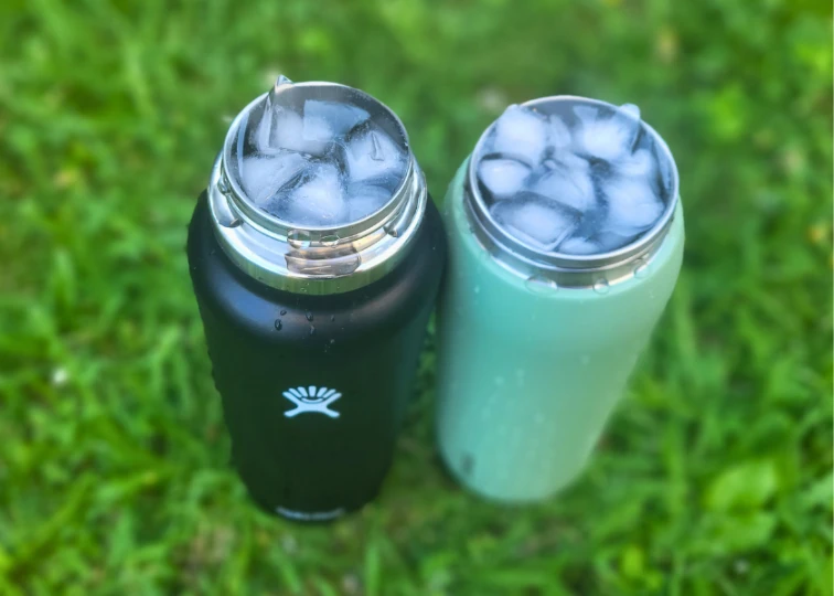 Two insulated water bottles, a black Hydro Flask and a mint green Owala FreeSip, filled with ice on grass.