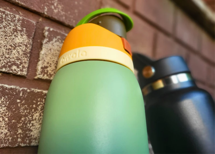 Close-up of two water bottle lids against a brick wall; a green Owala FreeSip and a black Hydro Flask.