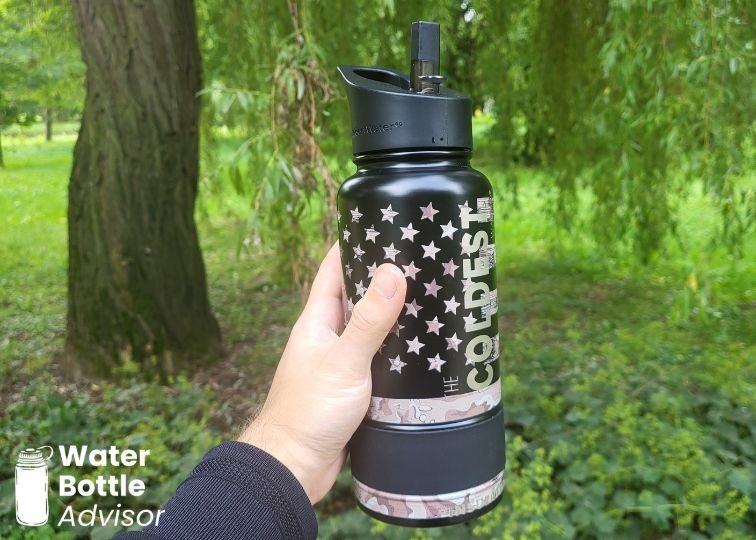 Coldest 32 oz black water bottle with flag-style design being held outdoors with straw lid open