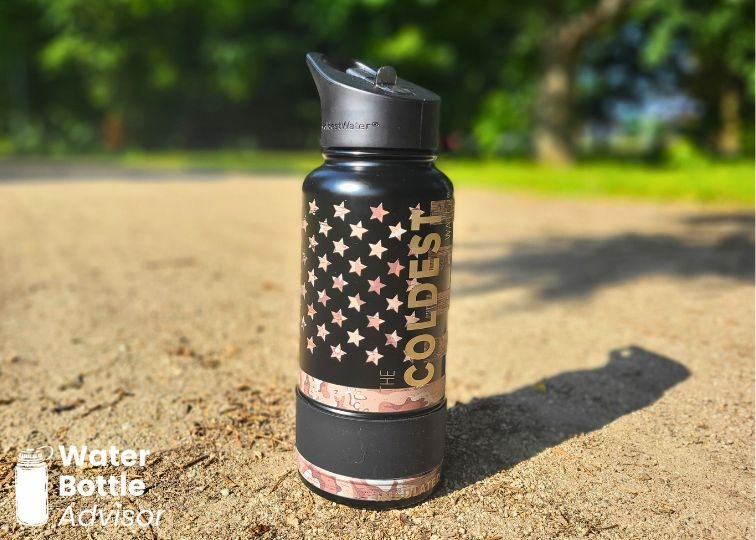 Coldest 32 oz black water bottle with flag-style design standing on a sunny outdoor path