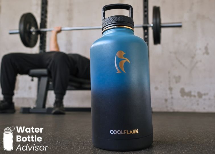 Coolflask 64 oz blue water bottle standing on a gym floor with workout equipment in the background
