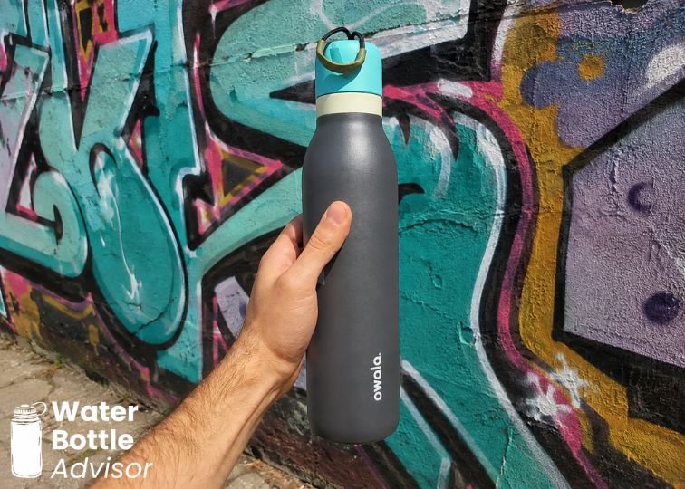 Owala FreeSip Twist 24 oz bottle being held outdoors in front of a colorful graffiti wall